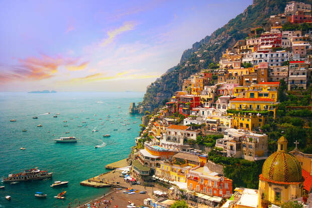 Colorful buildings cascade down a steep hillside against a backdrop of a calm blue sea with numerous boats, under a vivid, picturesque sky at sunset.